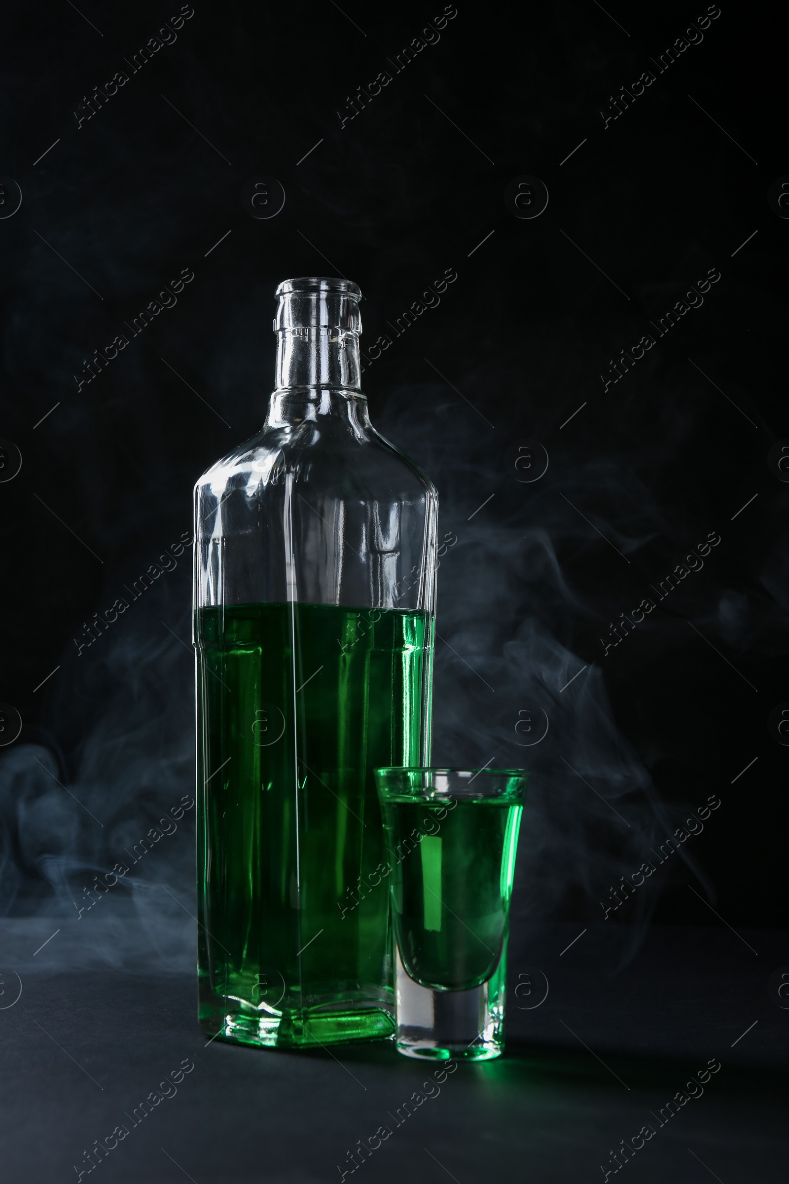 Photo of Absinthe in shot glass and bottle on table against black background with smoke