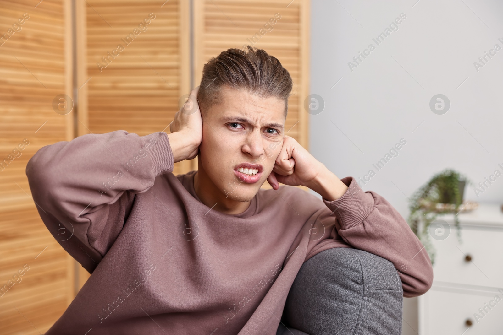 Photo of Annoyed young man covering his ears due to loud sound at home