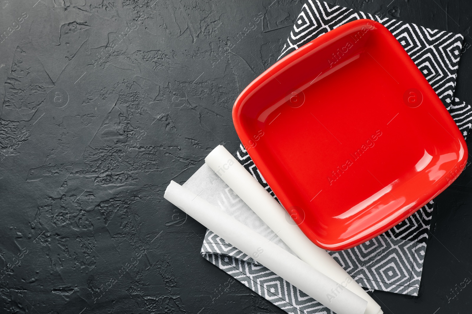 Photo of Red ceramic casserole and baking parchment paper on black table, top view. Space for text