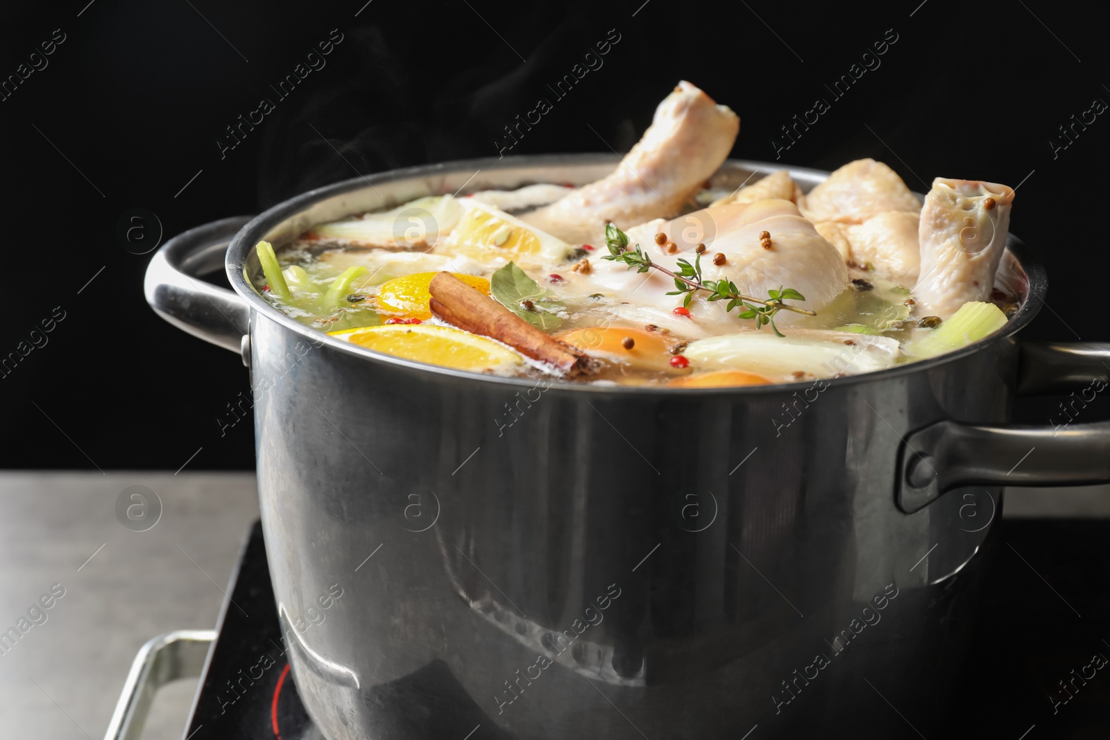 Photo of Whole fresh turkey, cut citrus fruits, thyme and spices boiling in pot on stove against black background, closeup