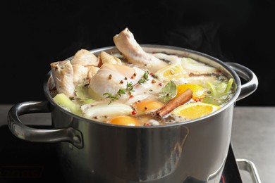 Photo of Whole fresh turkey, cut citrus fruits, thyme and spices boiling in pot on stove against black background, closeup