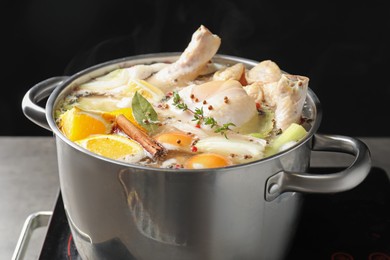 Photo of Whole fresh turkey, cut citrus fruits, thyme and spices boiling in pot on stove against black background, closeup