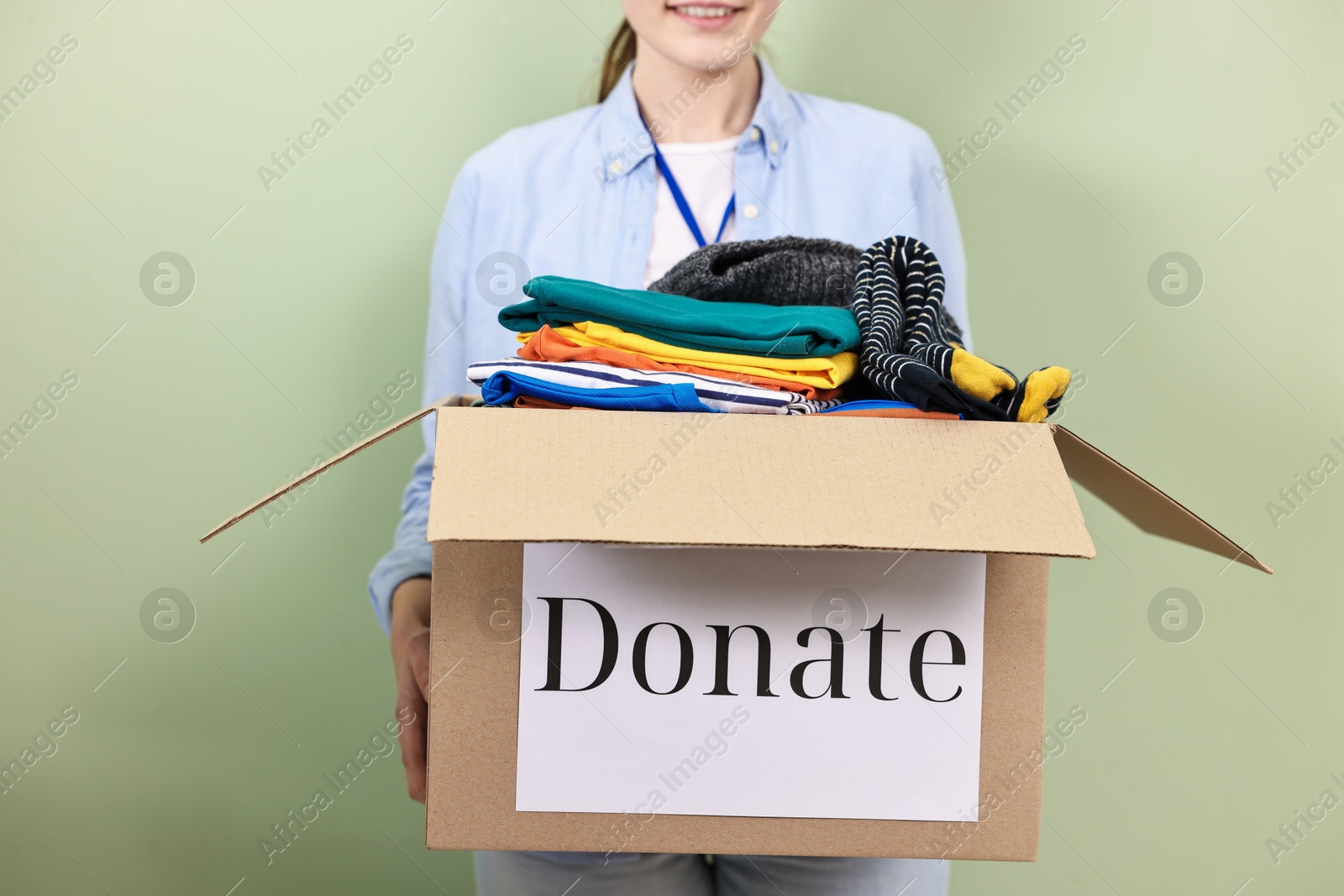 Photo of Woman holding donation box with clothes on pale olive background, closeup