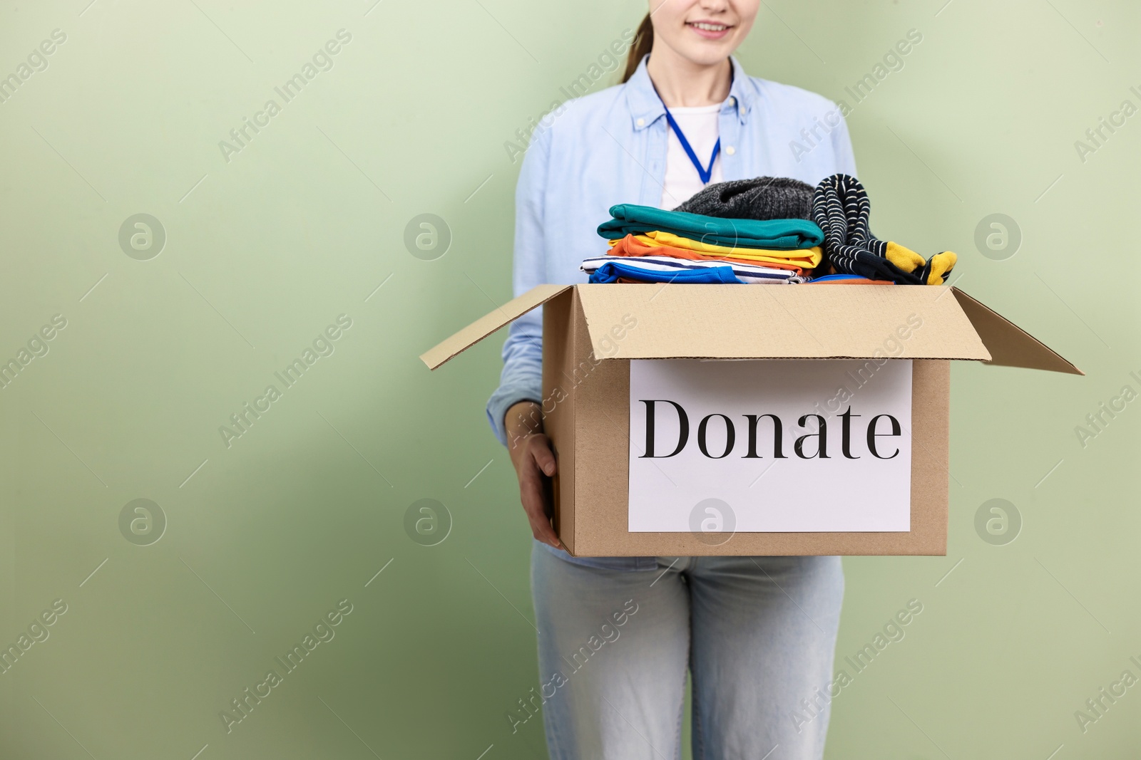 Photo of Woman holding donation box with clothes on pale olive background, closeup. Space for text