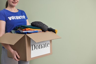 Photo of Woman holding donation box with clothes on pale olive background, closeup. Space for text