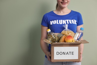 Photo of Woman holding donation box with clothes on pale olive background, closeup. Space for text