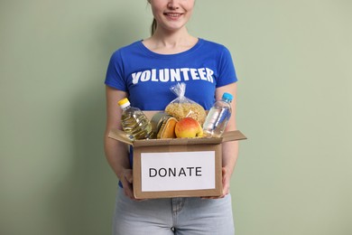 Photo of Woman holding donation box with food products on pale olive background, closeup