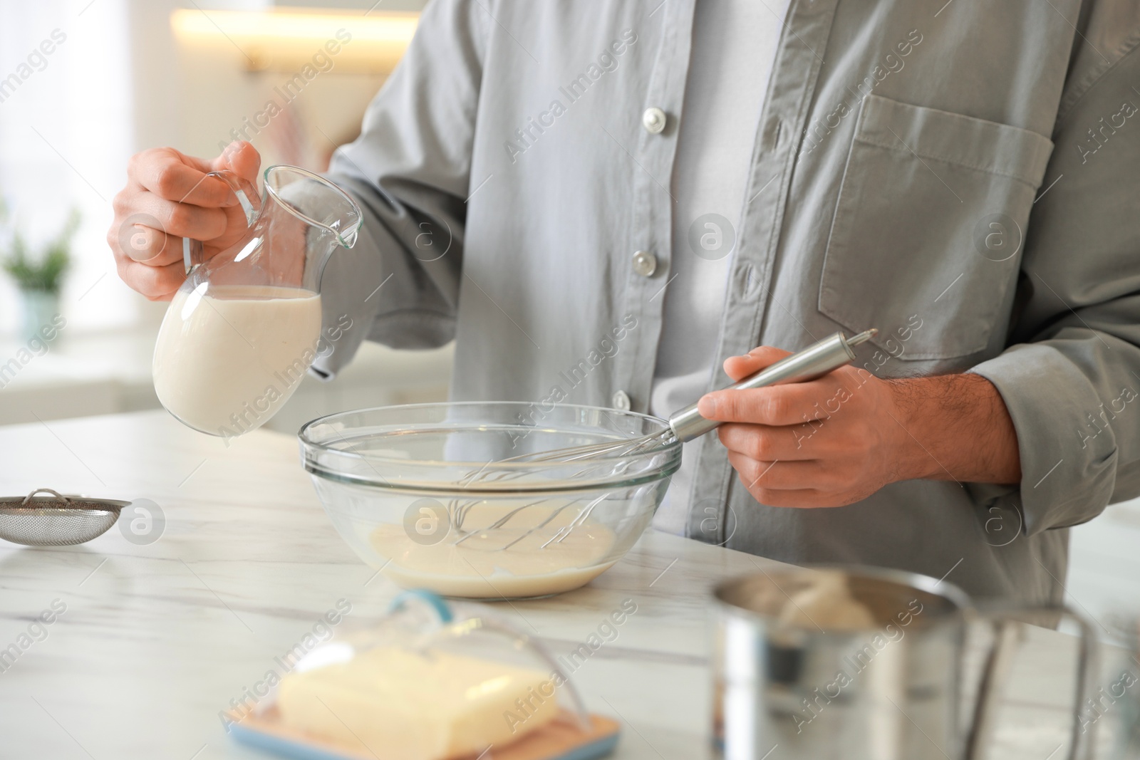 Photo of Making dough. Man pouring milk into bowl at white marble table in kitchen, closeup