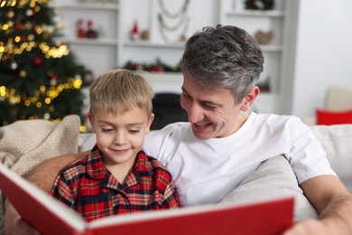 Photo of Father and son reading book together on sofa at home. Christmas holidays