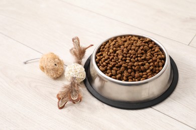 Photo of Dry pet food granules in feeding bowl and cat toys on floor indoors