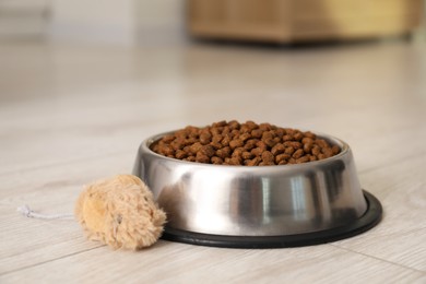 Photo of Dry pet food granules in feeding bowl and cat toy on floor indoors