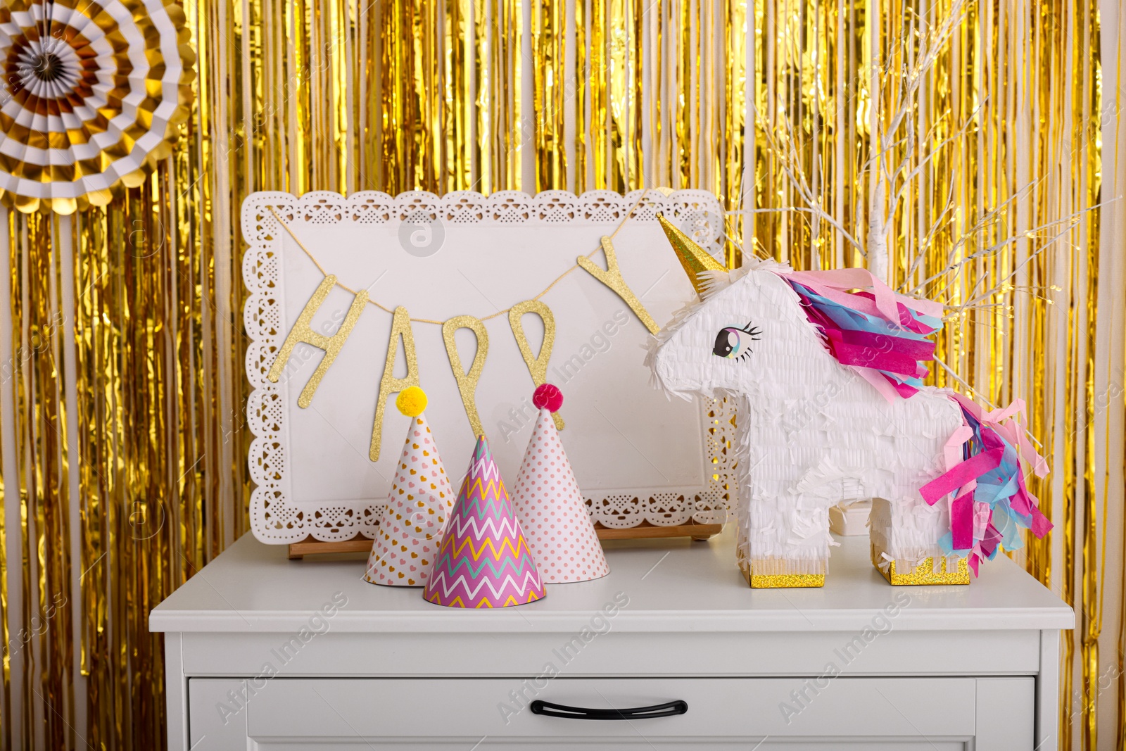 Photo of Bright pinata in shape of unicorn and party accessories on white chest of drawers against foil fringe curtain indoors