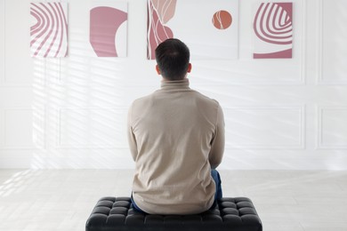 Photo of Man sitting on bench and looking at paintings in art gallery, back view