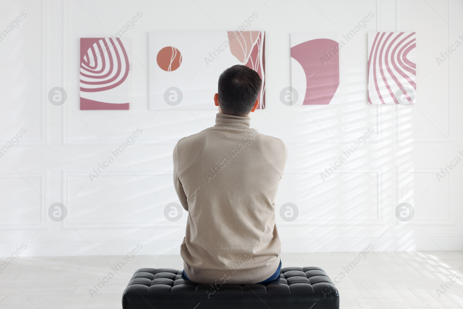 Photo of Man sitting on bench and looking at paintings in art gallery, back view