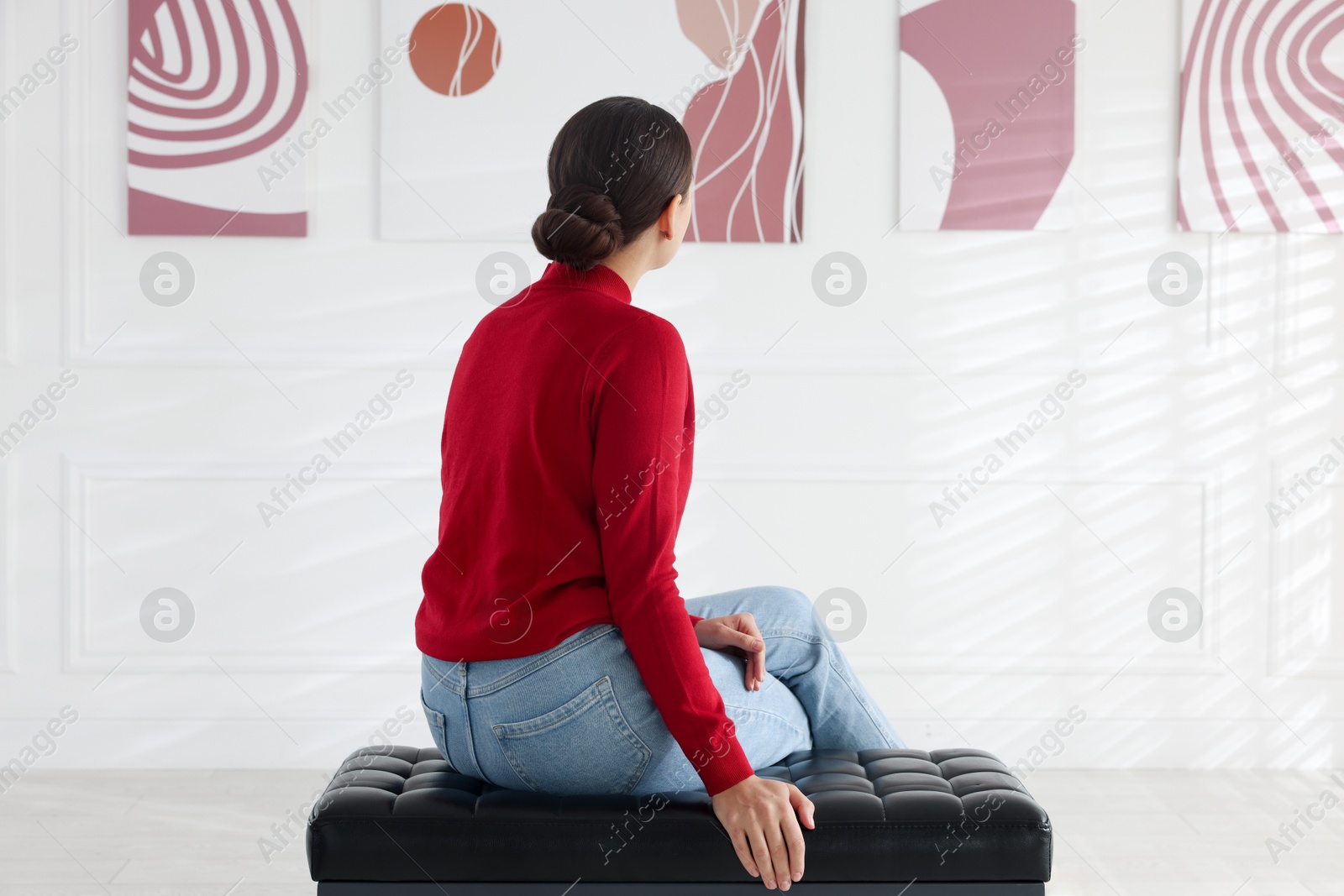 Photo of Woman sitting on bench and looking at paintings in art gallery, back view