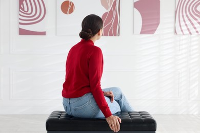 Photo of Woman sitting on bench and looking at paintings in art gallery, back view