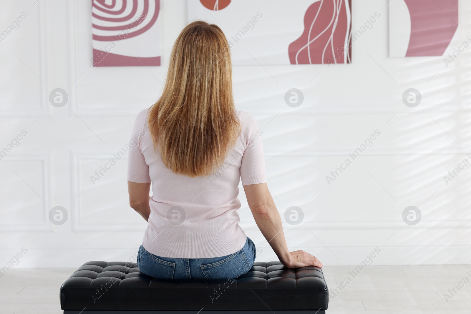 Photo of Woman sitting on bench and looking at paintings in art gallery, back view