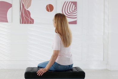 Photo of Woman sitting on bench and looking at paintings in art gallery, back view