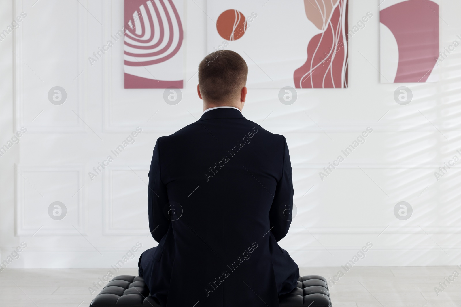 Photo of Man sitting on bench and looking at paintings in art gallery, back view