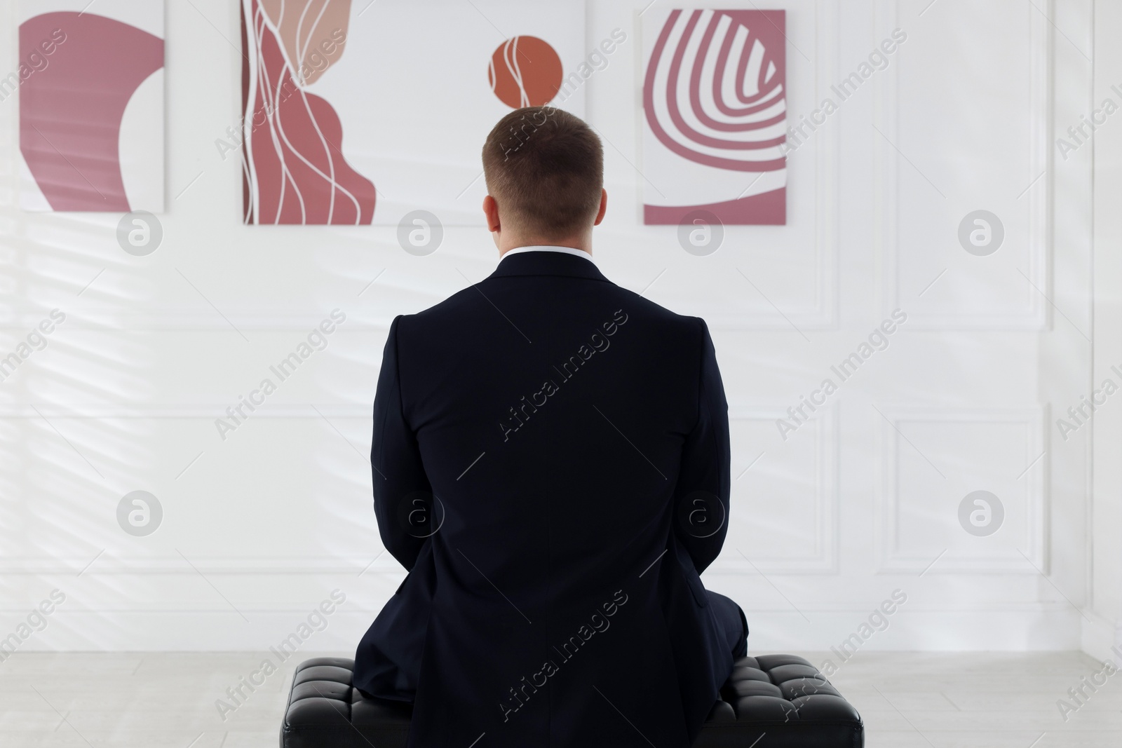 Photo of Man sitting on bench and looking at paintings in art gallery, back view