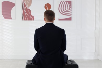 Photo of Man sitting on bench and looking at paintings in art gallery, back view