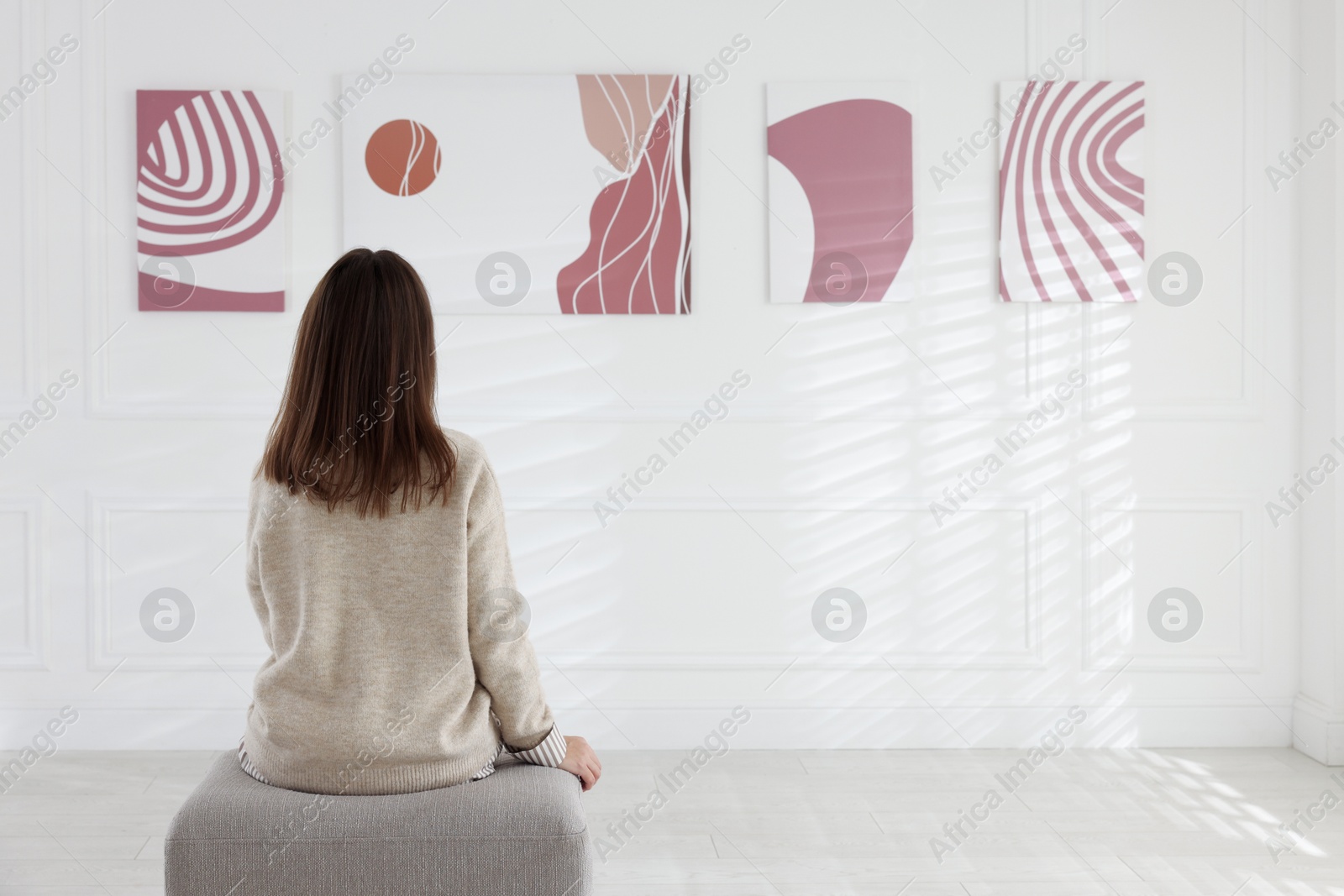 Photo of Woman sitting on pouf and looking at paintings in art gallery, back view