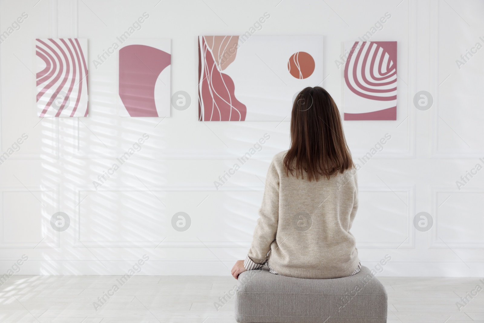 Photo of Woman sitting on pouf and looking at paintings in art gallery, back view