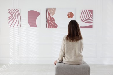 Photo of Woman sitting on pouf and looking at paintings in art gallery, back view