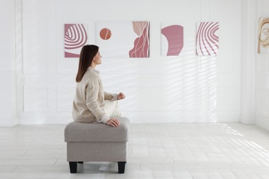 Photo of Woman sitting on pouf and looking at paintings in art gallery