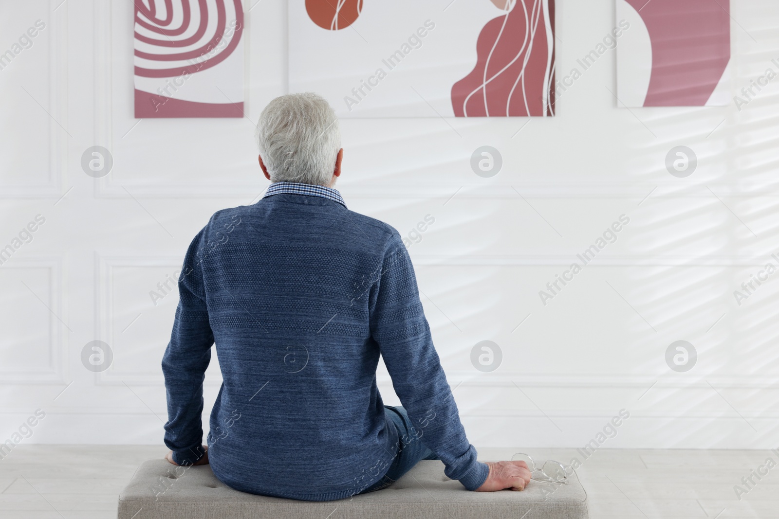 Photo of Senior man sitting on pouf and looking at paintings in art gallery, back view