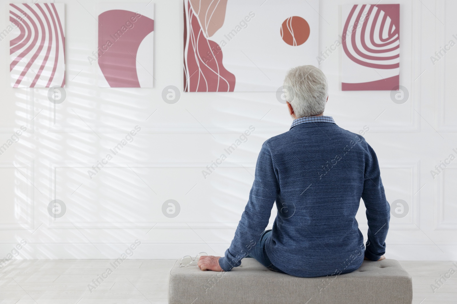 Photo of Senior man sitting on pouf and looking at paintings in art gallery, back view