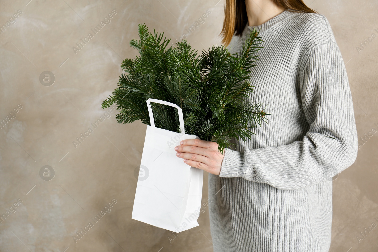 Photo of Woman holding green fir branches in paper bag near gray wall, closeup and space for text. Christmas decor