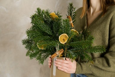 Photo of Woman holding beautiful Christmas composition of fir tree branches decorated with dried orange slices and cinnamon sticks near gray wall, closeup