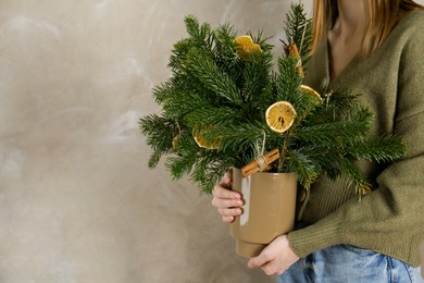 Photo of Woman holding beautiful Christmas composition of fir tree branches decorated with dried orange slices and cinnamon sticks near gray wall, closeup. Space for text