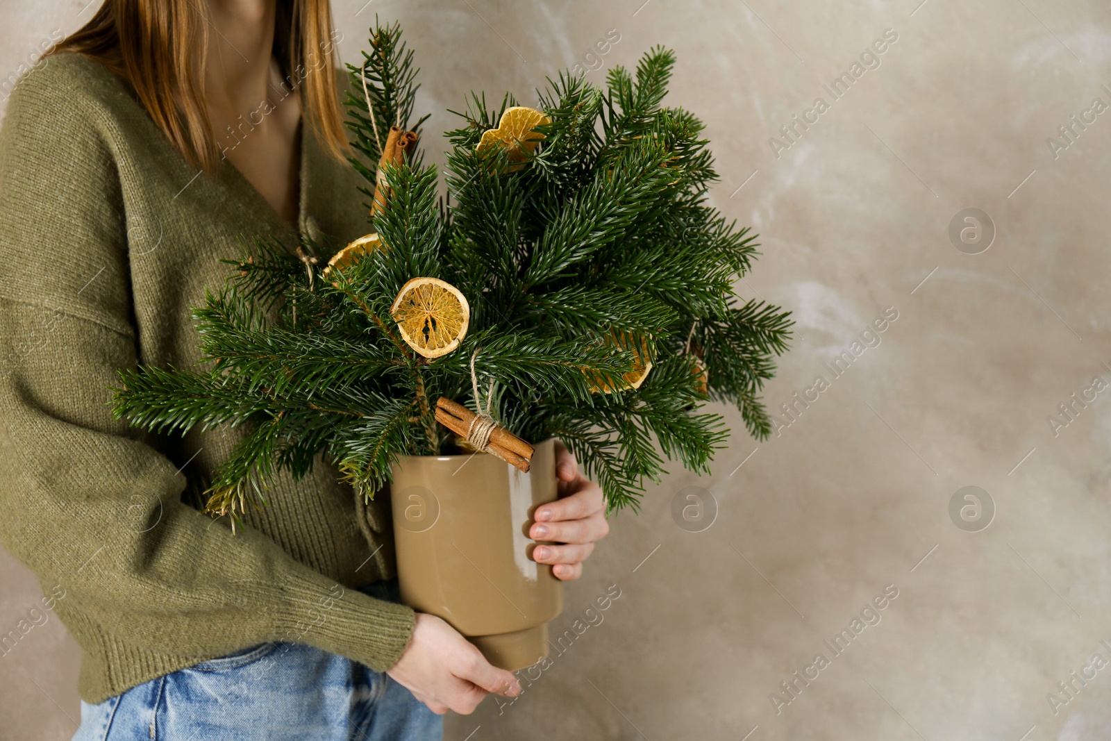 Photo of Woman holding beautiful Christmas composition of fir tree branches decorated with dried orange slices and cinnamon sticks near gray wall, closeup. Space for text