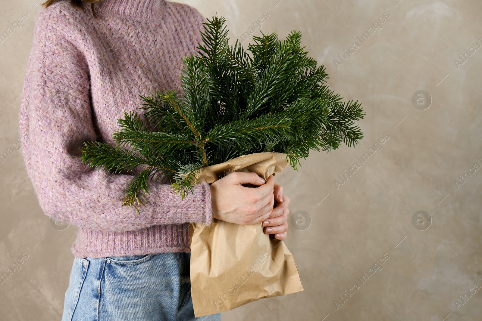 Photo of Woman holding green fir branches in paper bag near gray wall, closeup and space for text. Christmas decor