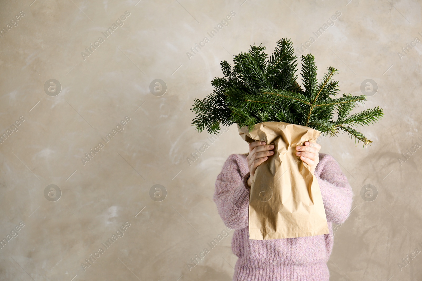 Photo of Woman holding green fir branches in paper bag near gray wall, space for text. Christmas decor