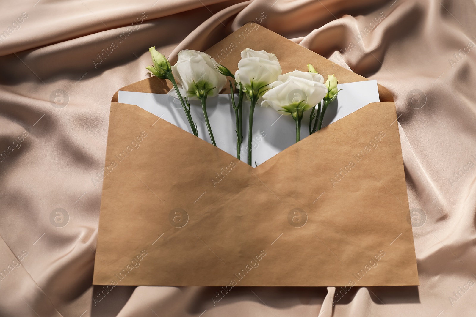 Photo of Kraft paper envelope with letter and flowers on beige silk fabric, above view. Mockup for design