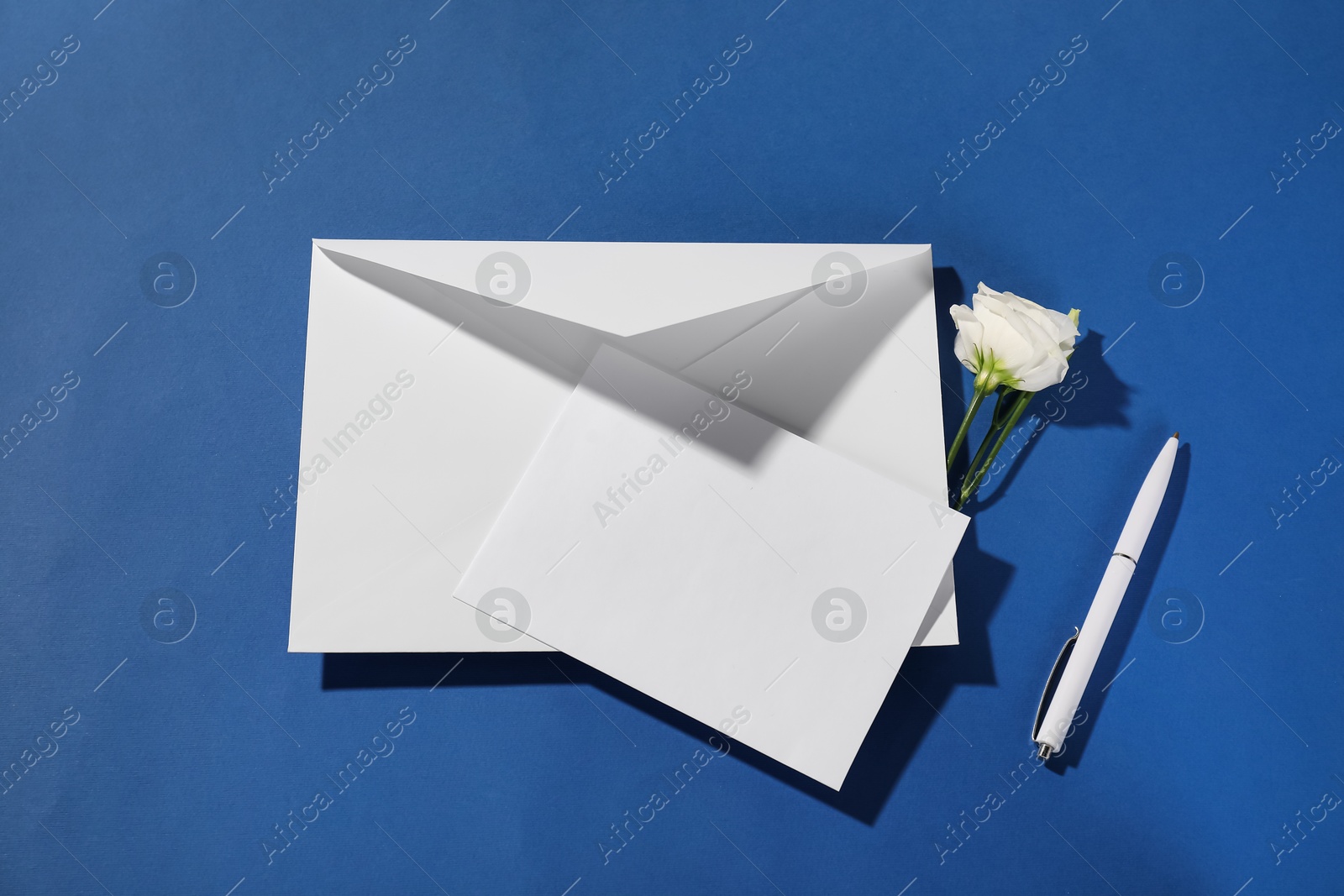 Photo of Paper envelope with letter, flower and pen on blue background, flat lay. Mockup for design