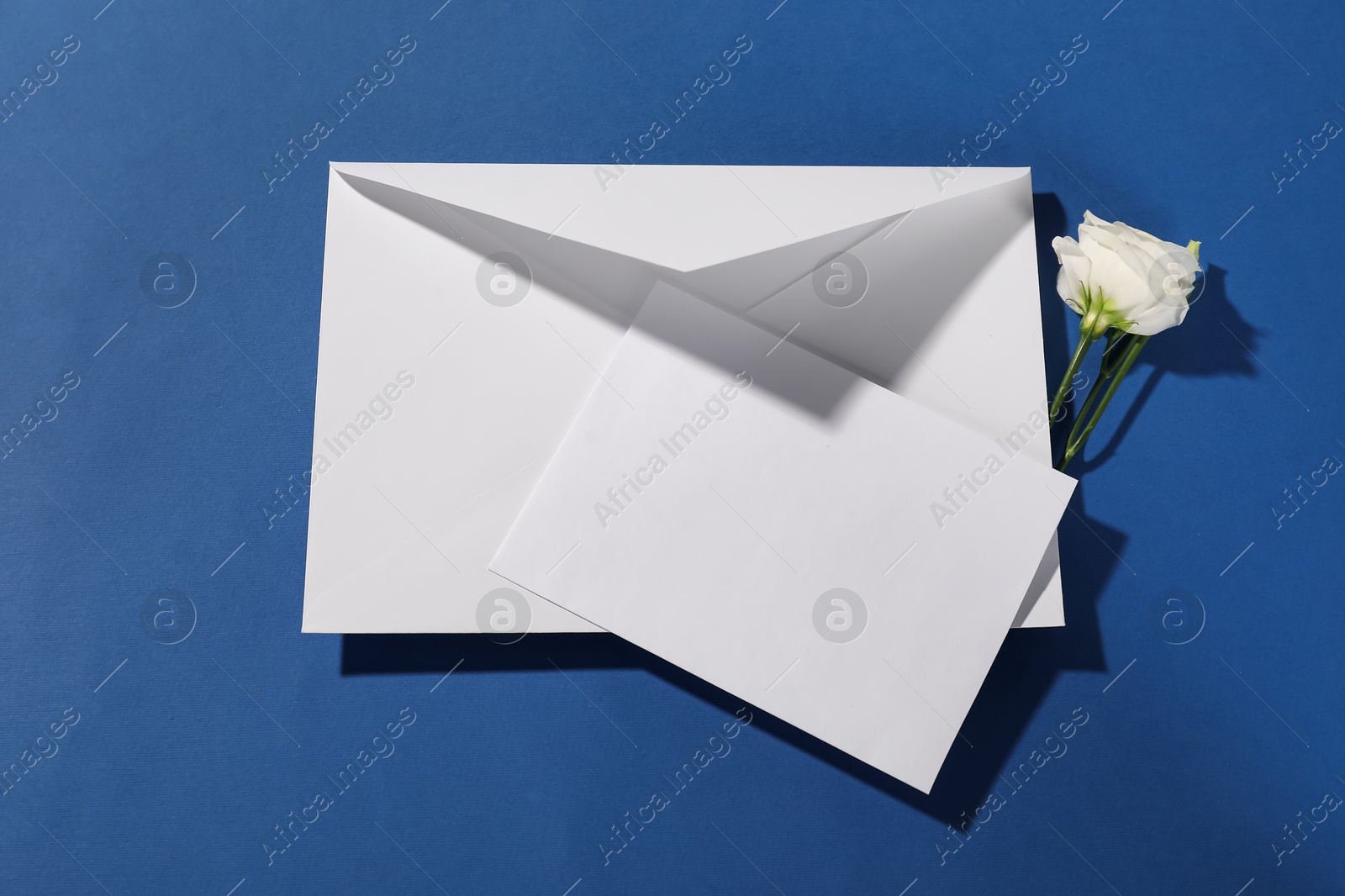 Photo of Paper envelope with letter and flower on blue background, top view. Mockup for design