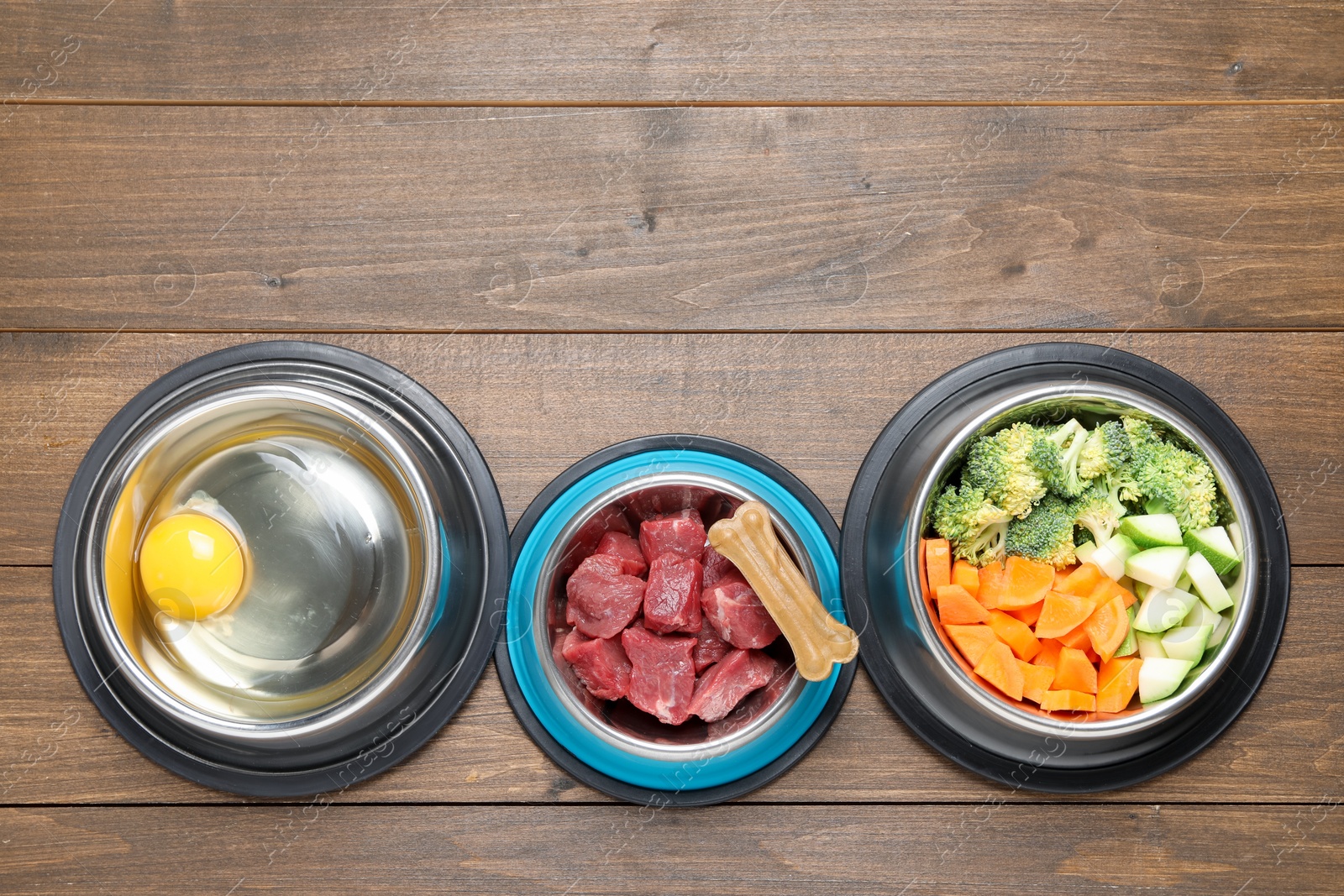 Photo of Natural pet food and chew bone in feeding bowls on wooden table, flat lay. Space for text