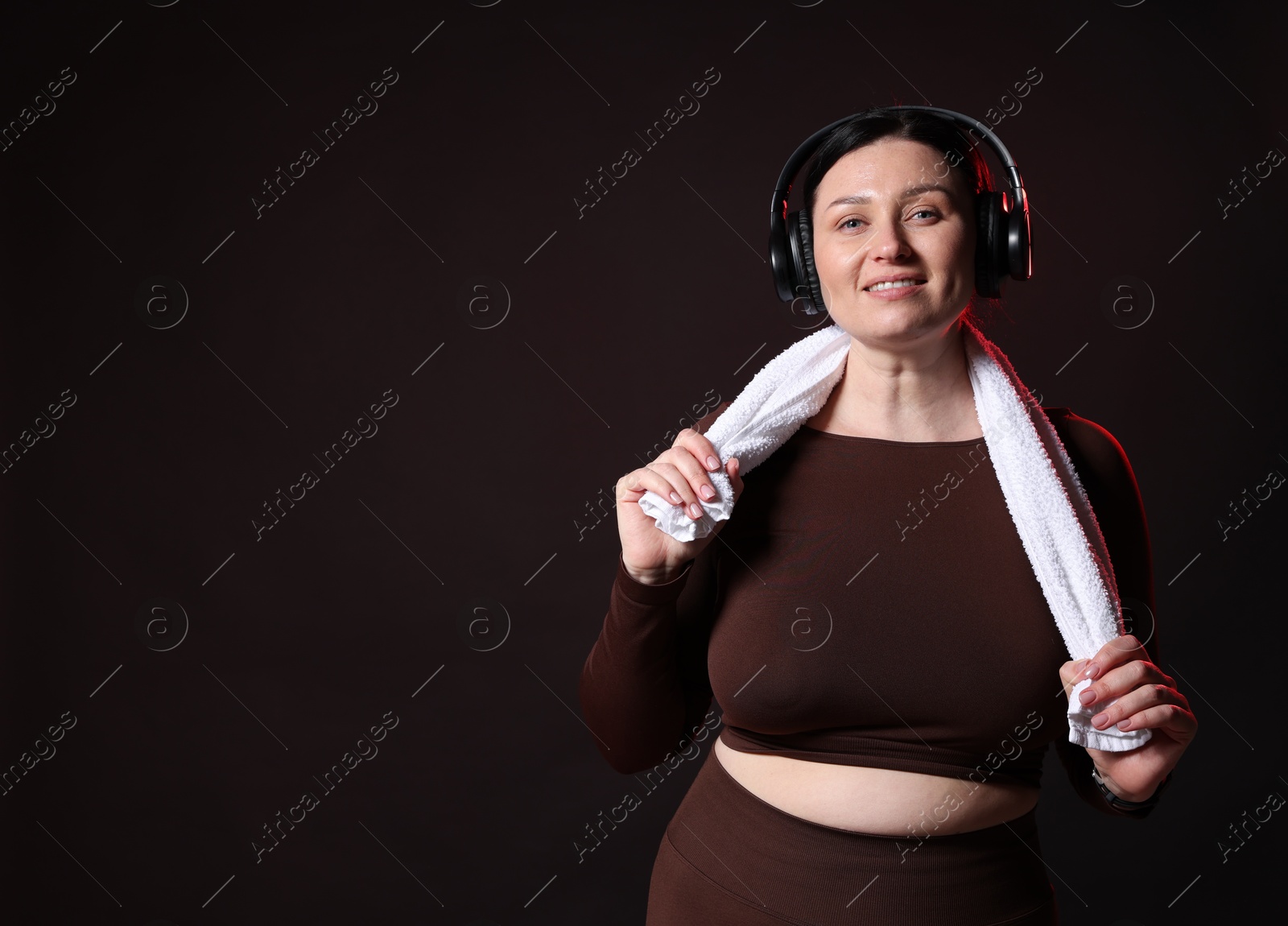 Photo of Plus size woman in gym clothes with headphones and towel on black background, space for text