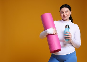 Photo of Plus size woman in gym clothes with water bottle and fitness mat on orange background, space for text