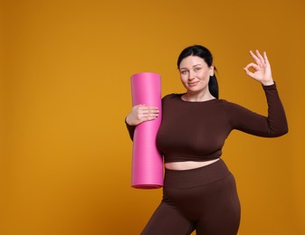 Photo of Plus size woman in gym clothes with fitness mat showing ok gesture on orange background, space for text