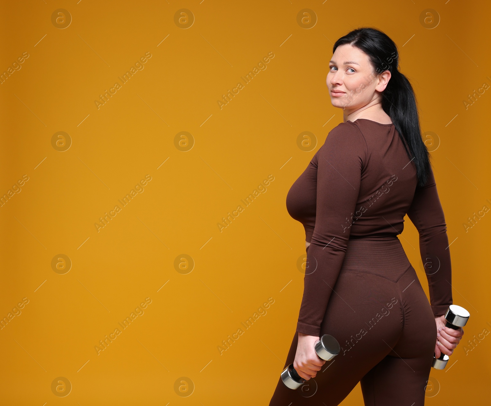 Photo of Plus size woman in gym clothes with dumbbells on orange background, space for text