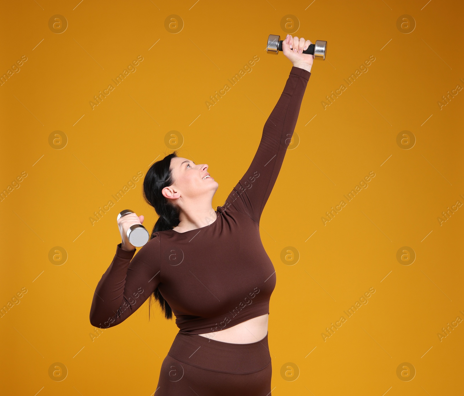 Photo of Plus size woman in gym clothes with dumbbells on orange background