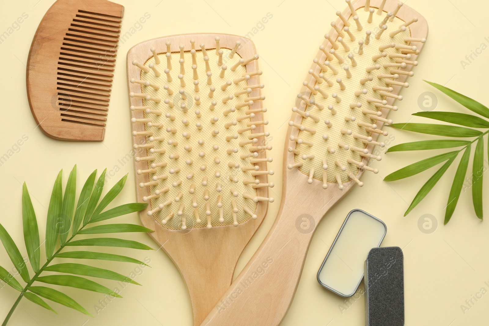 Photo of Wooden hair brushes, comb, wax and green leaves on beige background, flat lay