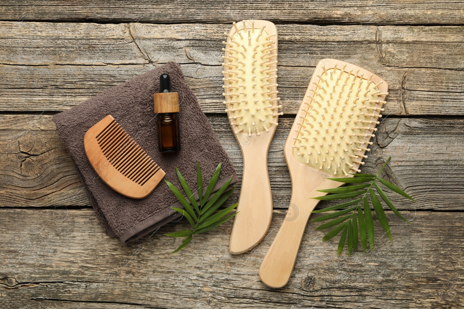 Photo of Hair brushes, comb, cosmetic product, towel and green leaves on wooden table, flat lay