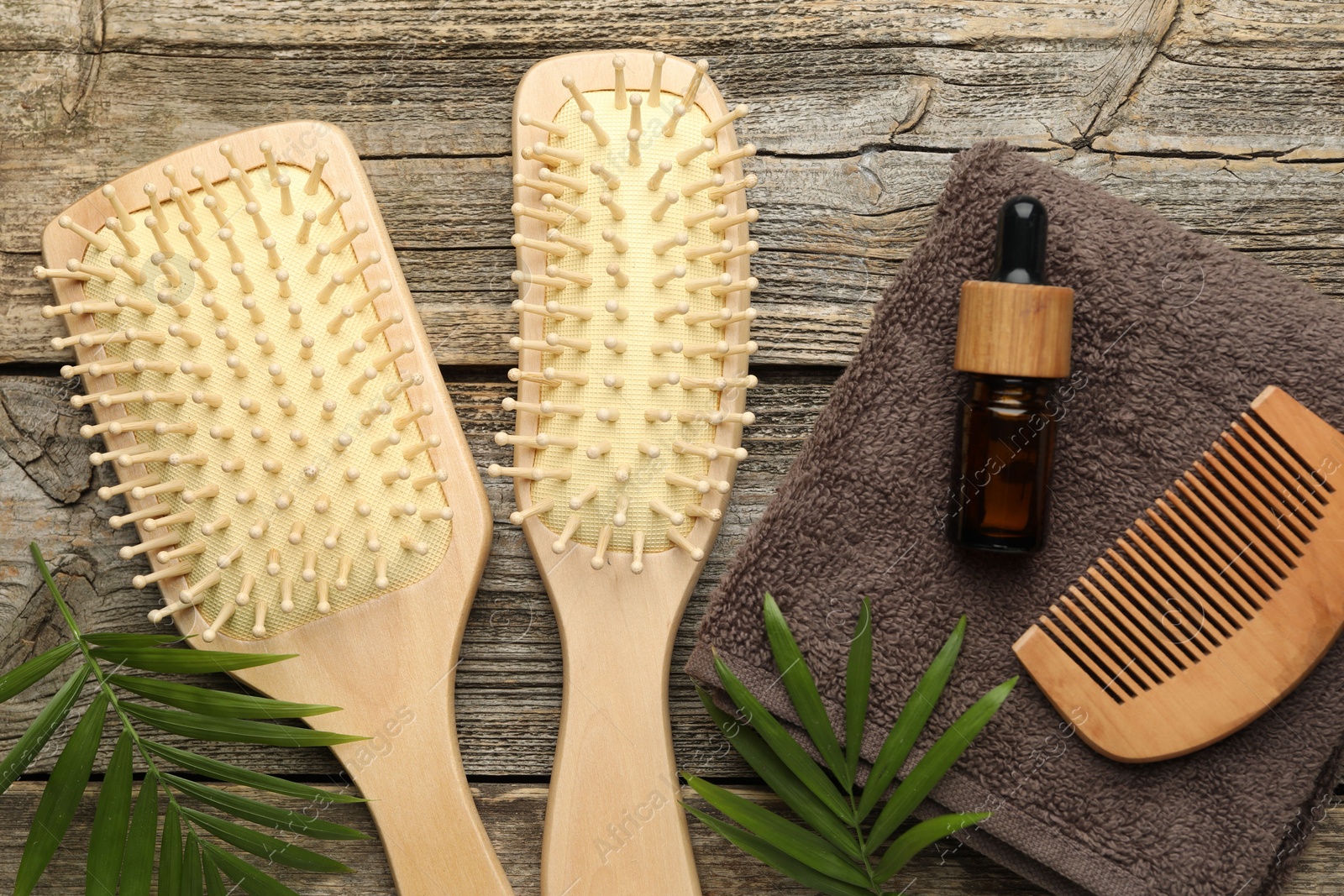 Photo of Hair brushes, comb, cosmetic product, towel and green leaves on wooden table, flat lay