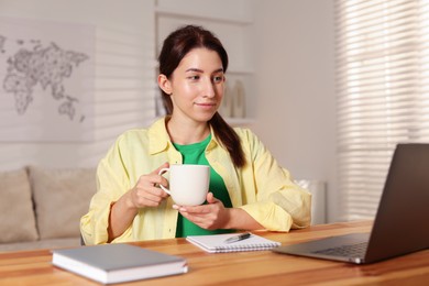 Photo of Young woman with cup of coffee working on laptop at desk in home office